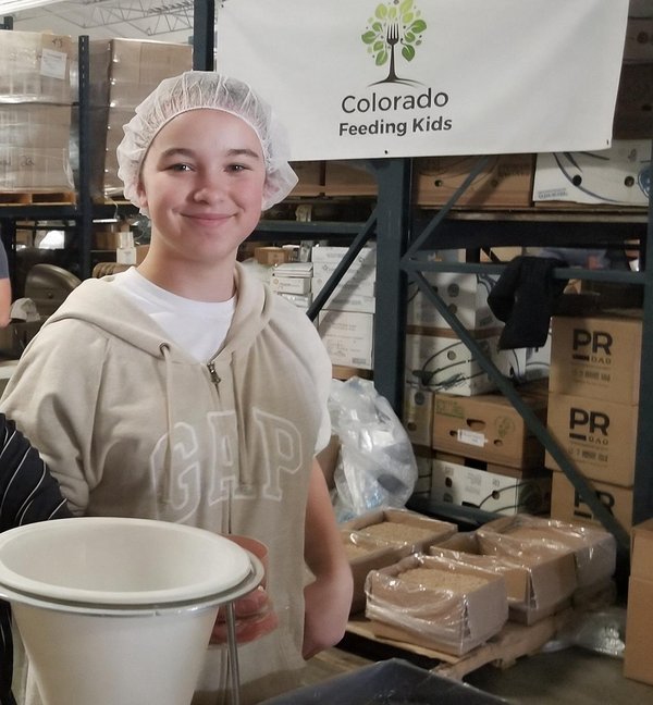 Teen volunteer packing meals, with the Colorado Feeding Kids sign in the background